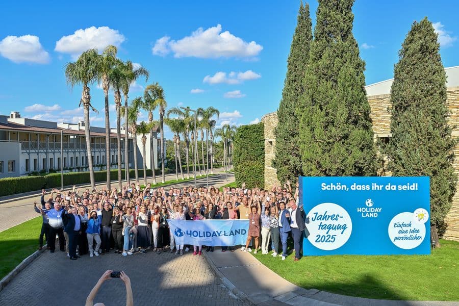 Gruppenfoto der HOLIDAY-LAND-Jahrestagung 2025 im Freien: Rund 200 Teilnehmende stehen lachend und winkend vor Palmen und Hotelgebäuden in der Lara Barut Collection in Antalya. Im Vordergrund hält eine Person ein HOLIDAY-LAND-Banner. Rechts steht ein großes blaues Willkommensschild mit den Logos von HOLIDAY LAND und dem Tagungsmotto „Einfach eine Idee persönlicher“. Der Himmel ist klar und blau.